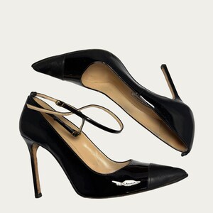 May include: Pair of black patent leather high heels with pointed toes and ankle straps. The heels have a stiletto design and a small silver emblem on the side. The shoes are set against a neutral background.