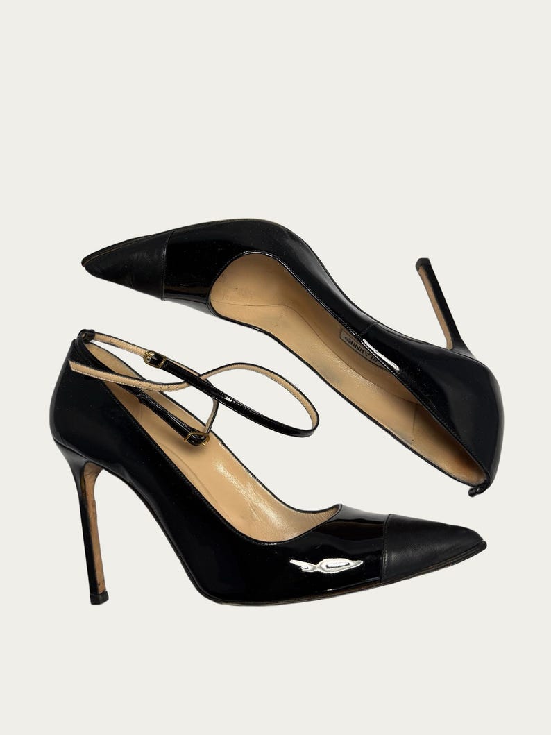 May include: Pair of black patent leather high heels with pointed toes and ankle straps. The heels have a stiletto design and a small silver emblem on the side. The shoes are set against a neutral background.