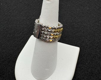 Puede incluir: Un anillo de cuentas plateadas y doradas con un cierre rectangular plateado. El anillo se exhibe en un expositor de joyas blanco en forma de dedo sobre un fondo negro. El anillo tiene múltiples filas de pequeñas cuentas.