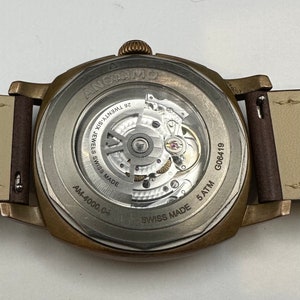 May include: Close-up of the back of a bronze-colored ANONIMO watch with a transparent window showcasing the intricate inner workings. The watch has a brown leather strap and the text "26 TWENTY-SIX JEWELS SWISS MADE" is visible.