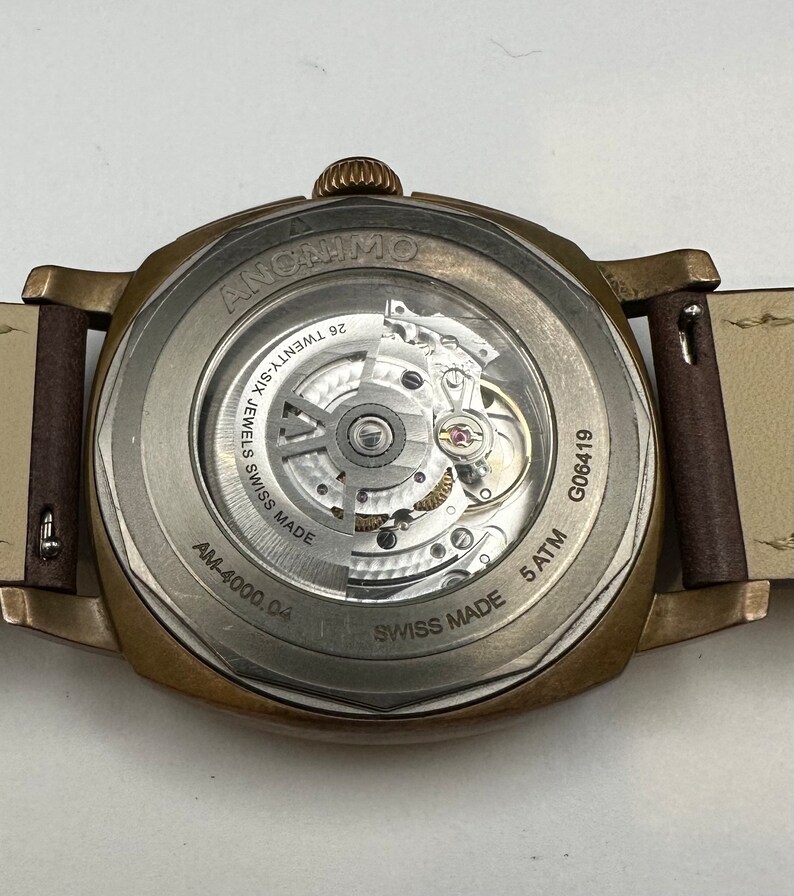 May include: Close-up of the back of a bronze-colored ANONIMO watch with a transparent window showcasing the intricate inner workings. The watch has a brown leather strap and the text "26 TWENTY-SIX JEWELS SWISS MADE" is visible.