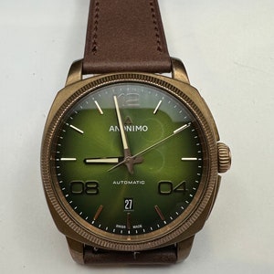 May include: A bronze-colored wristwatch with a green face and brown leather strap. The watch face displays the brand name "ANONIMO", the word "AUTOMATIC", and the date "27". The watch has a round face with large numerals at the 4, 8, and 12 o'clock positions.