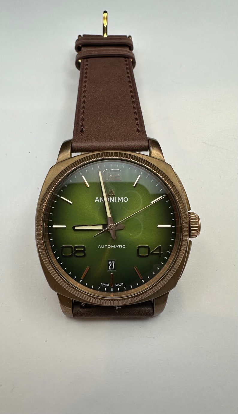 May include: A bronze-colored wristwatch with a green face and brown leather strap. The watch face displays the brand name "ANONIMO", the word "AUTOMATIC", and the date "27". The watch has a round face with large numerals at the 4, 8, and 12 o'clock positions.