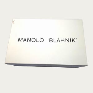 May include: A white rectangular box with the brand name "MANOLO BLAHNIK" printed in black. The box is slightly angled, with a few minor imperfections on the surface.