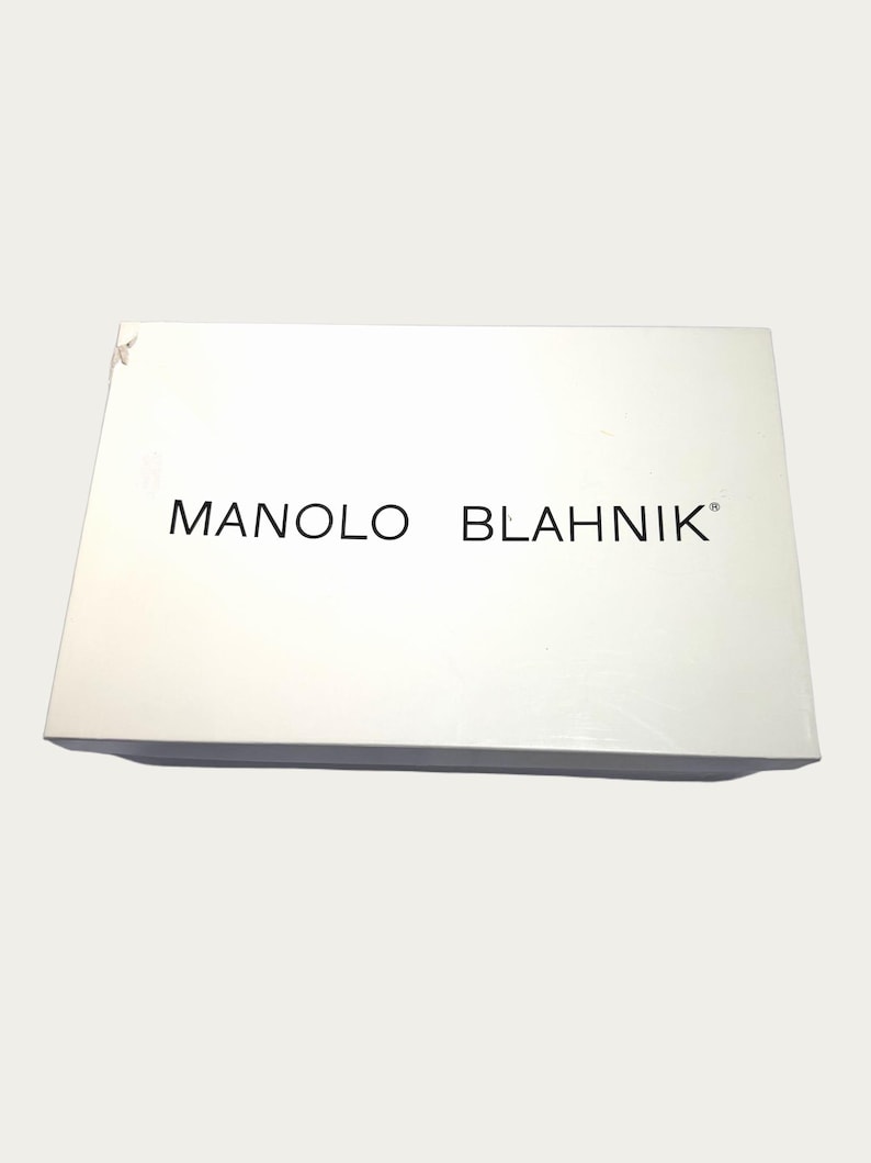 May include: A white rectangular box with the brand name "MANOLO BLAHNIK" printed in black. The box is slightly angled, with a few minor imperfections on the surface.