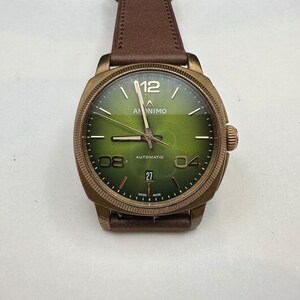 May include: A bronze-toned wristwatch with a green face and brown leather strap. The watch face displays the brand name "ANONIMO", the word "AUTOMATIC", and the date. The watch has a textured bezel and a brown leather strap.