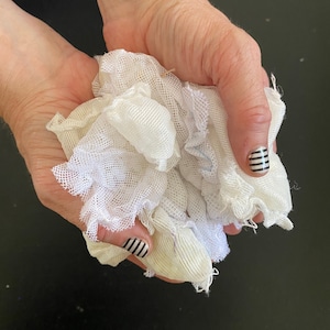 Könnte beinhalten: Eine Handvoll weißen Gazestoffs, zusammengeknüllt. Der Stoff hat ein zartes, offenes Gewebe und einen leicht gerüschten Rand. Die Hände, die den Stoff halten, haben manikürte Nägel mit schwarz-weiß gestreiften Designs.