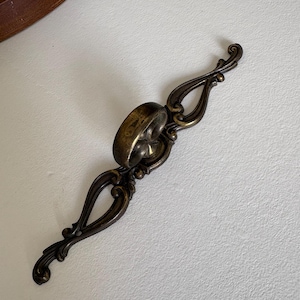 May include: An antique bronze-toned drawer pull with an ornate, symmetrical design. The pull features a central ring and decorative, curved arms with scroll-like details. The hardware is likely for a cabinet or drawer.