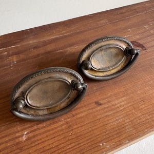 May include: Two antique brass drawer pulls with an oval design. The pulls have a decorative beaded edge and a curved handle. The brass has a tarnished, aged appearance, adding to their vintage charm. They are set against a wooden surface.