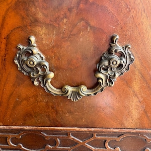 May include: Close-up of a wooden cabinet door with a decorative brass handle. The wood has a rich, reddish-brown color with visible grain patterns. The handle is ornate, with a curved design and intricate detailing, suggesting an antique or vintage style.
