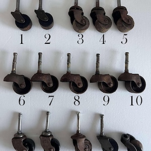 May include: Fifteen antique furniture casters are arranged in rows. The casters have a variety of wheel designs, with metal stems and varying degrees of rust. Numbers 1 through 15 are printed below each caster.