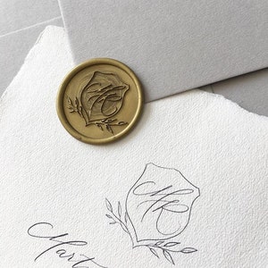 Peut inclure: Un sceau de cire doré avec un monogramme et un motif de feuilles, posé sur un papier blanc texturé. Le sceau est rond et présente les initiales "MK" dans une écriture élégante. Le papier a un bord déchiré, et le fond comprend du papier gris.