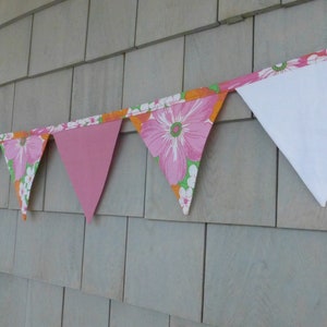 Peut inclure: Une guirlande de fanions rose et blanche à bordure orange est accrochée à un mur en bois gris. La guirlande est en tissu et a un bord festonné.