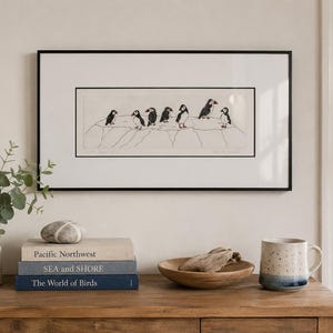 May include: A framed horizontal print featuring a line drawing of seven puffins standing on a rocky cliff edge. The artwork is enclosed in a thin black frame with a white mat, displaying a detailed illustration of the birds in black and white.
