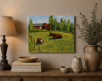 May include: An oil painting depicts two brown cows resting in a green pasture with a red barn in the background. The artwork is displayed on a wooden surface with a lamp, books, and decorative pottery.
