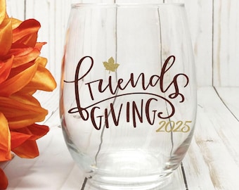 May include: A clear stemless wine glass with the words "FriendsGIVING 2025" in a brown script font. A brown leaf graphic is above the word "GIVING". An orange and yellow flower is on the left side of the glass.