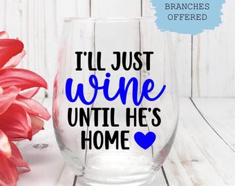 May include: A clear stemless wine glass with the text "I'll just wine until he's home" in black and blue. A blue heart is at the bottom. A large red flower is on the left side of the image.