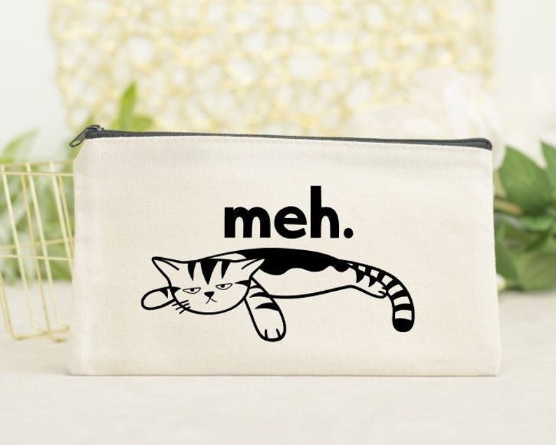 May include: A white canvas makeup bag with a black and white illustration of a cat with the text "meh." printed on it.