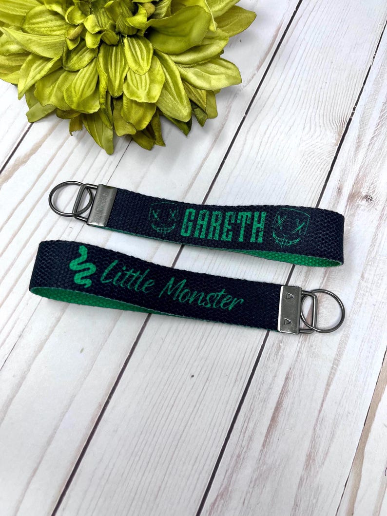 Op de afbeelding: Twee sleutelhangers met een marineblauwe buitenkant en een groene binnenkant. Op de ene sleutelhanger staat "Gareth" in groene letters met een afbeelding. Op de andere sleutelhanger staat "Little Monster" in groene letters met een slangafbeelding. Elke sleutelhanger heeft een zilverkleurige metalen ring.