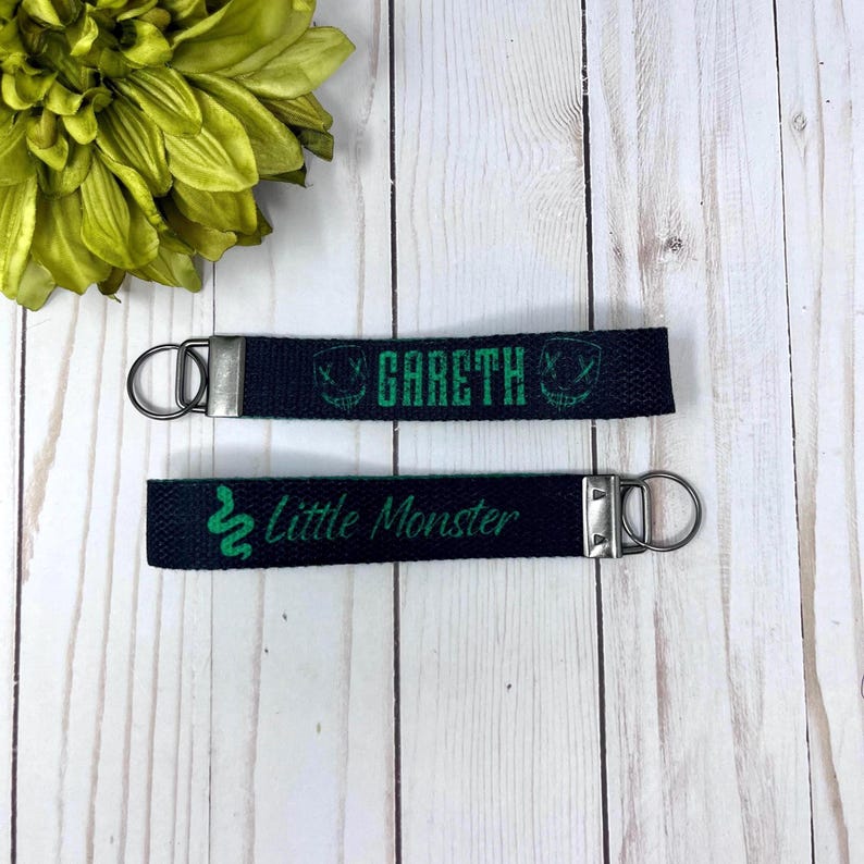 Op de afbeelding: Twee zwarte sleutelhangers met zilverkleurige hardware. E&eacute;n sleutelhanger heeft de naam "Gareth" in groen, met een groene afbeelding. De andere sleutelhanger zegt "Little Monster" in groen met een groene slangafbeelding.