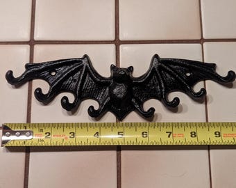 Op de afbeelding: Zwarte vleermuisvormige wandhaak van gietijzer. De vleermuis heeft uitgespreide vleugels en drie gebogen haken. De spanwijdte is ongeveer 25 cm. Perfect voor Halloween of gotische decoratie.