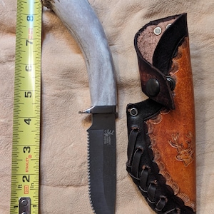 May include: A hunting knife with a serrated blade and a handle made from an antler. The knife is next to a leather sheath with a deer design. The blade is approximately 4 inches long.