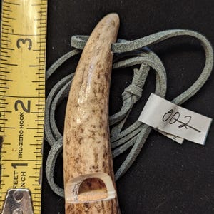 Può includere: Un fischietto artigianale in corno di cervo con un colore marrone e crema screziato naturale. Il fischietto ha una camera sonora scolpita ed è attaccato a un cordino di cuoio grigio. L'etichetta recita "002". Il fischietto è lungo circa 7.6 cm.