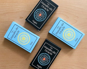 Puede incluir: Cuatro cartas de oráculo, dos azules y dos negras, con el texto "The Hanabi-shi Oracle" y "Day" o "Night" impreso en cada carta. Las cartas presentan un diseño circular con un patrón central en forma de flor.