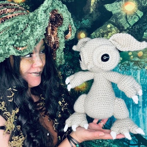 Puede incluir: Una mujer con un sombrero de crochet verde y marr&oacute;n sostiene una criatura gris de crochet con grandes garras blancas. La criatura tiene grandes ojos negros y una peque&ntilde;a boca. El fondo es una escena de bosque verde y marr&oacute;n.