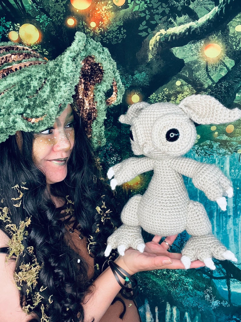 Puede incluir: Una mujer con un sombrero de crochet verde y marr&oacute;n sostiene una criatura gris de crochet con grandes garras blancas. La criatura tiene grandes ojos negros y una peque&ntilde;a boca. El fondo es una escena de bosque verde y marr&oacute;n.