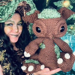 Puede incluir: Una mujer con un sombrero de crochet verde y marr&oacute;n sostiene una criatura de crochet marr&oacute;n y verde con ojos grandes. La criatura tiene garras blancas y est&aacute; sentada en la mano de la mujer.