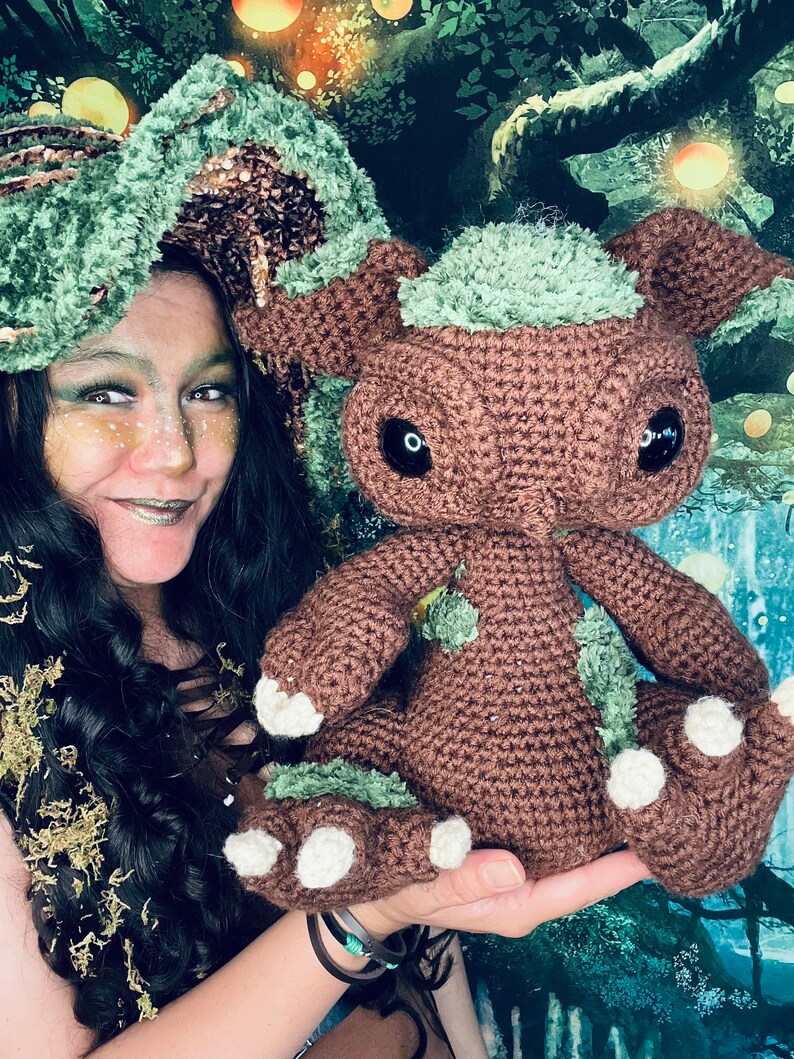 Puede incluir: Una mujer con un sombrero de crochet verde y marr&oacute;n sostiene una criatura de crochet marr&oacute;n y verde con ojos grandes. La criatura tiene garras blancas y est&aacute; sentada en la mano de la mujer.