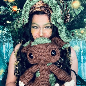 Puede incluir: Una persona que lleva un gorro de crochet verde y marr&oacute;n sostiene un juguete de peluche de crochet marr&oacute;n con detalles verdes. El juguete tiene ojos negros grandes y una expresi&oacute;n amigable.
