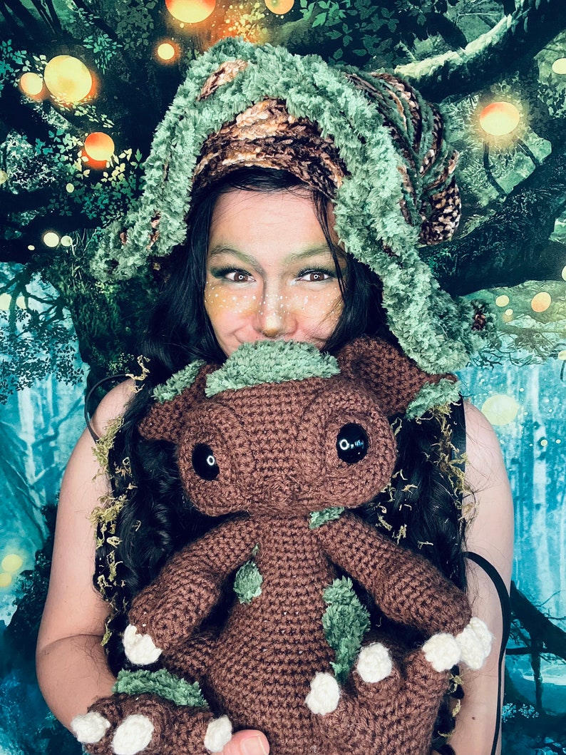 Puede incluir: Una persona que lleva un gorro de crochet verde y marr&oacute;n sostiene un juguete de peluche de crochet marr&oacute;n con detalles verdes. El juguete tiene ojos negros grandes y una expresi&oacute;n amigable.