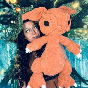 Puede incluir: Una mujer con un sombrero verde y un vestido marr&oacute;n sostiene un gran juguete de peluche naranja con grandes ojos negros. El juguete tiene orejas largas y peludas y garras blancas. El fondo es una imagen borrosa de un bosque con luces brillantes.