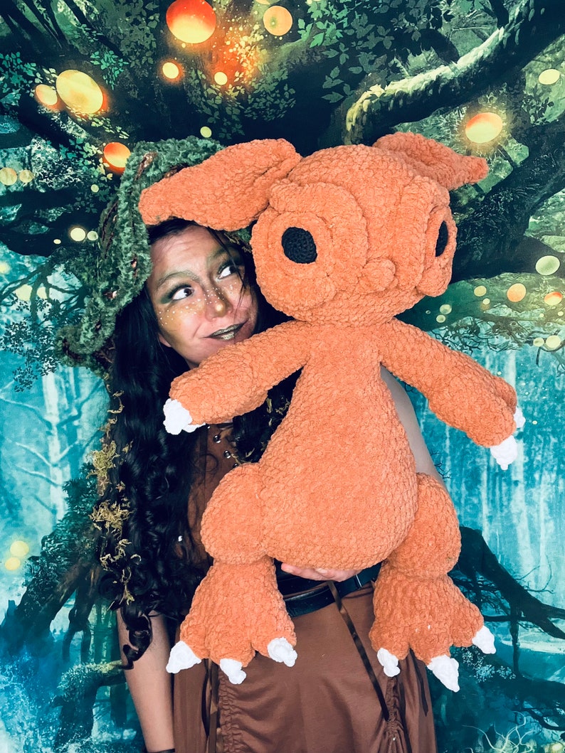Puede incluir: Una mujer con un sombrero verde y un vestido marr&oacute;n sostiene un gran juguete de peluche naranja con grandes ojos negros. El juguete tiene orejas largas y peludas y garras blancas. El fondo es una imagen borrosa de un bosque con luces brillantes.