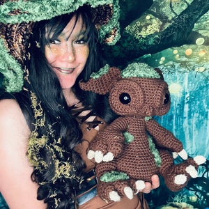 Puede incluir: Una mujer con un sombrero de crochet verde y marr&oacute;n sostiene un juguete de peluche de crochet marr&oacute;n y verde. El juguete tiene ojos grandes y una expresi&oacute;n amigable. La mujer y el juguete est&aacute;n frente a un fondo verde y azul con una escena de bosque.