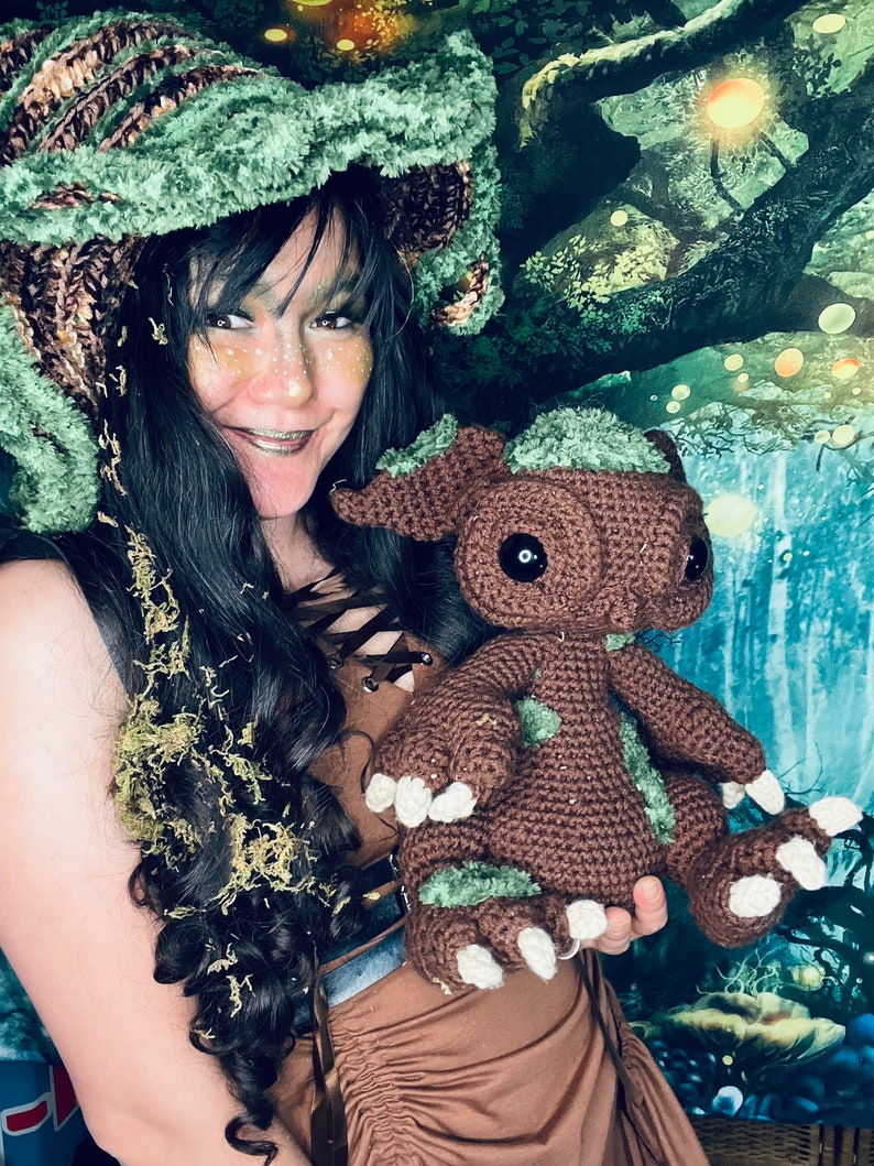 Puede incluir: Una mujer con un sombrero de crochet verde y marr&oacute;n sostiene un juguete de peluche de crochet marr&oacute;n y verde. El juguete tiene ojos grandes y una expresi&oacute;n amigable. La mujer y el juguete est&aacute;n frente a un fondo verde y azul con una escena de bosque.