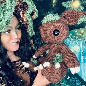 Puede incluir: Una mujer con un sombrero de crochet verde y marr&oacute;n sostiene un juguete de peluche de crochet marr&oacute;n con detalles verdes. El juguete tiene un ojo negro grande y pies blancos. El fondo es una escena de bosque verde y marr&oacute;n.