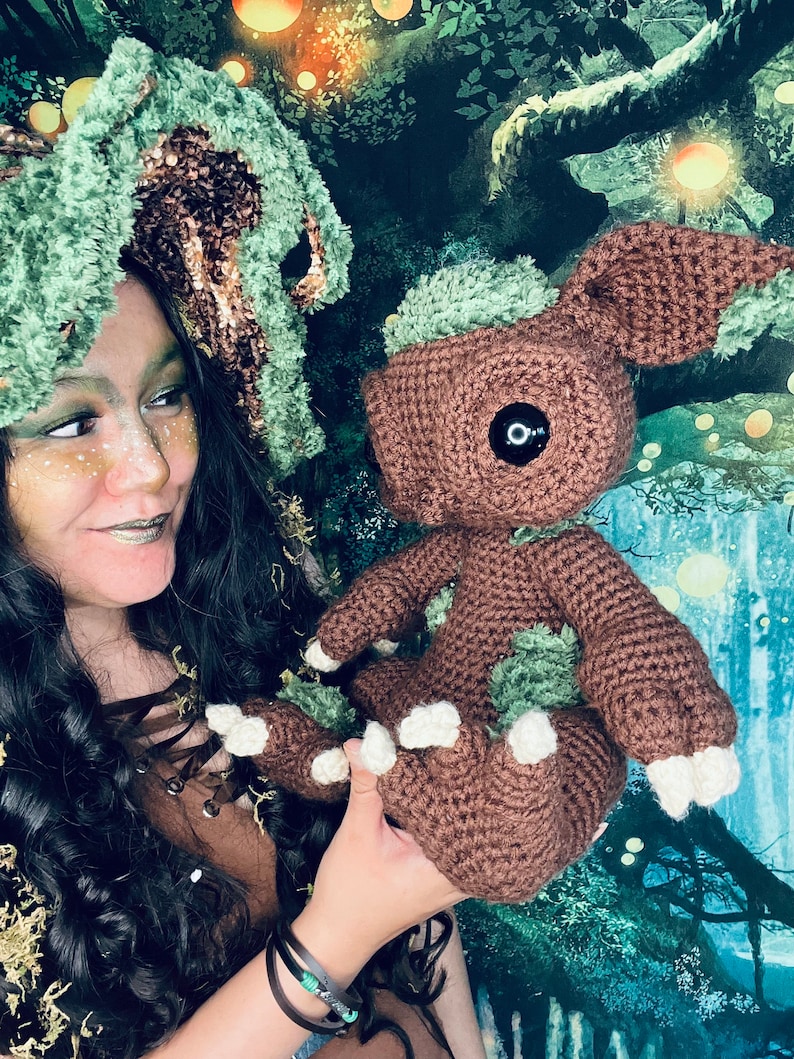 Puede incluir: Una mujer con un sombrero de crochet verde y marr&oacute;n sostiene un juguete de peluche de crochet marr&oacute;n con detalles verdes. El juguete tiene un ojo negro grande y pies blancos. El fondo es una escena de bosque verde y marr&oacute;n.