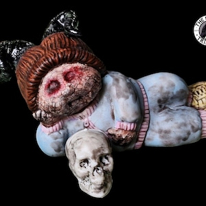 Op de afbeelding: Een griezelige, blauw getinte pop met hoorns en een schedel in zijn hand. De pop heeft een gebarsten, geschilderd gezicht met rode ogen en een roze-wit gestreept shirt. De pop ligt op zijn zij met gebogen benen en voeten in gele schoenen. De pop is gelabeld met "The Ravens Trinkets".