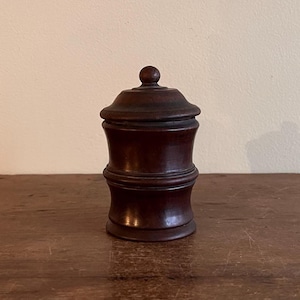 May include: A small, dark brown wooden container with a lid. The container has a rounded, hourglass shape with a small knob on top. The wood grain is visible, and the piece appears to be antique or vintage.