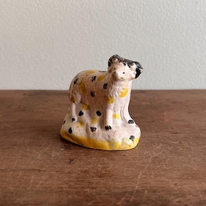 May include: A small, vintage ceramic sheep figurine. The sheep is painted with a cream base, yellow accents, and black spots. The sheep stands on a yellow and cream base. The sheep's head and ears are black.