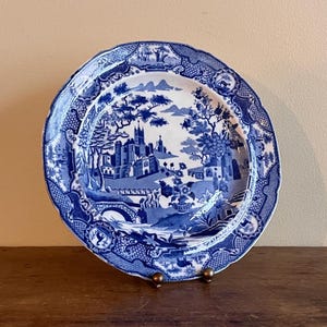 Puede incluir: Plato de cerámica azul y blanco con una escena de paisaje detallada. El plato presenta un borde festoneado y un diseño clásico. El paisaje incluye edificios, árboles y un puente.