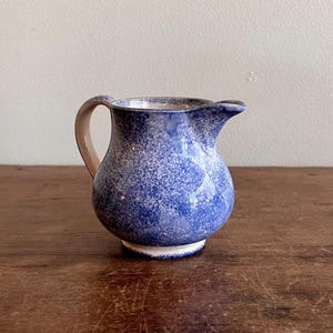 May include: A small ceramic jug with a speckled blue glaze. The jug has a rounded body, a curved handle, and a spout for pouring. The base is white, and the handle is a light tan colour. The jug is on a wooden surface.