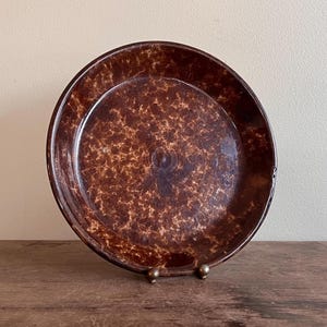 May include: A brown speckled plate with a raised rim. The plate is displayed on a small metal stand. The plate's surface has a mottled pattern of light and dark brown.