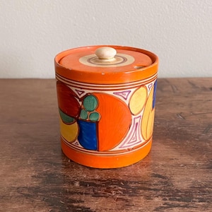 May include: A cylindrical orange container with a lid and a small wooden knob. The container is decorated with colorful geometric shapes in blue, yellow, red, and green. The lid is orange with a light wood center.