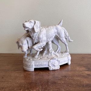 May include: A white ceramic figurine depicting two dogs. One dog stands, while the other is positioned lower, with its head near the first dog's legs. The base features decorative relief patterns and a small clock design.