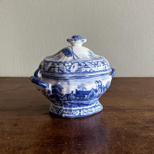 Könnte beinhalten: Eine blau-weiße Zuckerdose aus Keramik mit Deckel, verziert mit einem detaillierten Transferdruck-Muster aus ländlichen Landschaften, Häusern und Bäumen. Die Dose hat zwei seitliche Griffe und einen kleinen Knauf am Deckel sowie ein florales Randmuster.