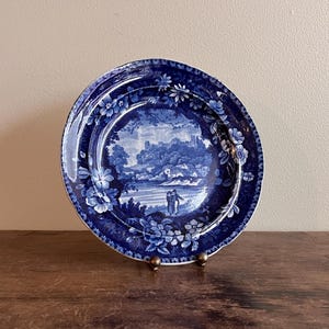 May include: A blue and white decorative plate with a landscape scene. The plate features a detailed illustration of a river, trees, and figures. Floral patterns adorn the rim. The plate is displayed on a wooden surface.